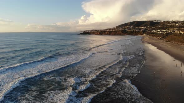 A drone flies high above crashing waves at the beach with a beautiful sky backdrop.