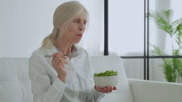 Mature Whitehaired Woman Sits on the Couch and Eats a Vegetable Salad