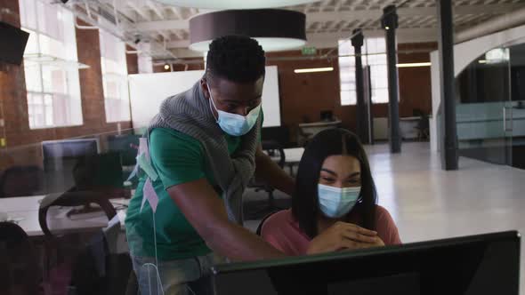 Diverse male and female business colleagues wearing face masks in discussion at work using computer