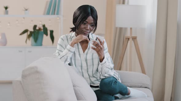 Attractive Young Woman Sitting on Comfortable Sofa in Cozy Living Room Holding Mobile Phone Relaxed