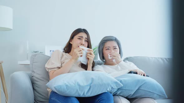 Asian senior mature woman and daughter sitting on sofa watch sad movie on TV and cry together.