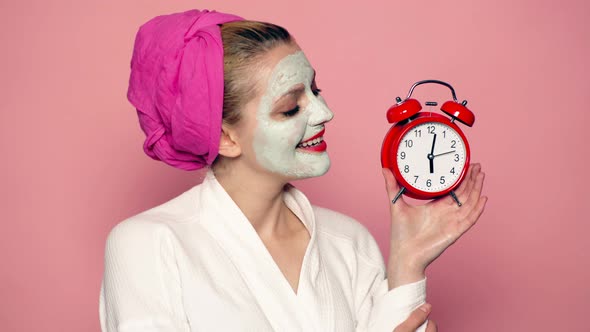 Funny Young Woman with Alarm Clock at Morning. Girl with a Mask on His Face Holds a Clock on a Pink