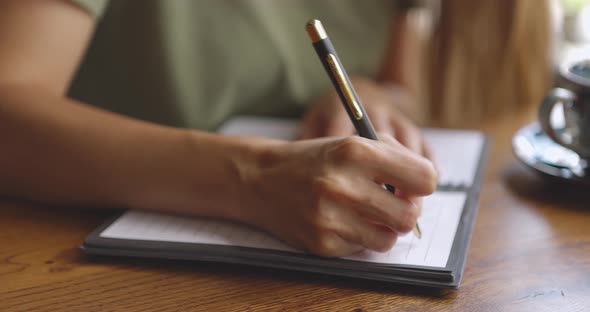 Close Up of Woman's Hands Writing Down on a White Blank Notebook with Cup on Wooden Table