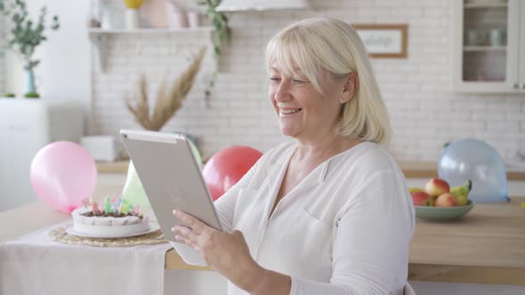 Positive Mature Woman Scrolling Tablet Screen and Smiling While Standing in the Modern Kitchen