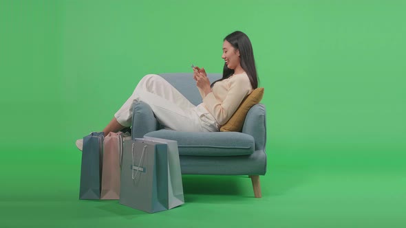 Woman With Shopping Bags Using Mobile Phone Shopping Online While Sitting On Sofa At Green Screen