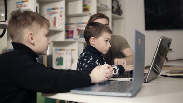 Two Young Boys are Learning How to Use Laptops with Help and Guidance of Young Programmer Teacher