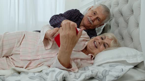 Senior Elderly Couple Wearing Pyjamas Lying on Bed Looking on Mobile Phone Laughing and Having Fun