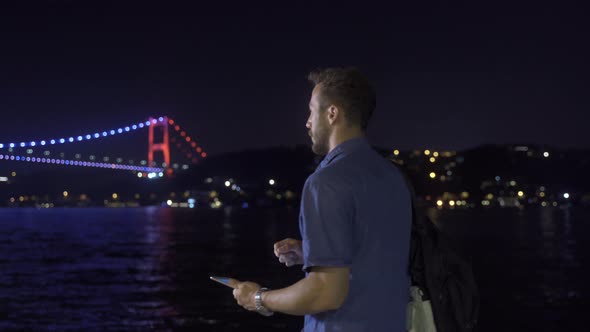 Man using tablet near illuminated bridge in strait.