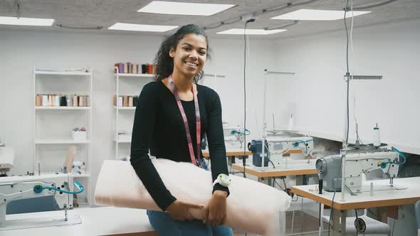 Mixed Race Lady Tailor Holding Roll of Tulle Fabric and Smiling Posing Sitting on Workstation