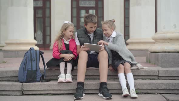 Wide Shot of Three Classmates Sitting on Break Outdoors and Surfing Internet on Tablet. Caucasian