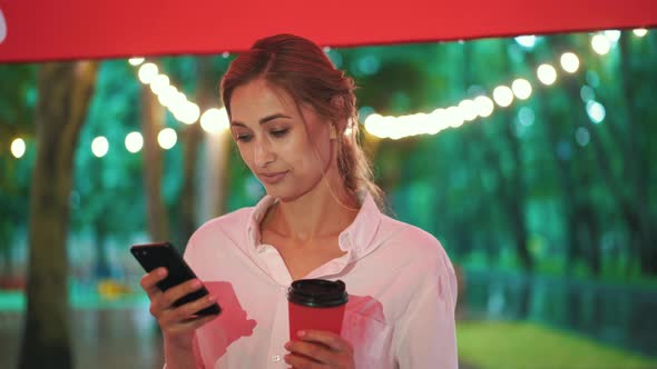 Woman Stands Under Canopy During The Rain In City Park Use Phone Holds Coffee In Hand