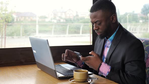 Black Businessman Works on Laptop with Smartphone in Cafe and Drinks Coffee