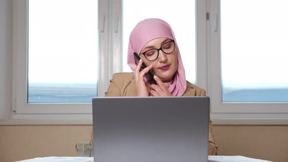 Woman in Hijab Examines Documents and Works on Laptop While Talking on the Phone