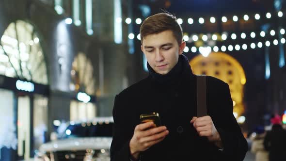 A Good-looking Man in a Coat Walks Down the Street Using a Smartphone