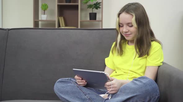 The Little One Smiles and Looks at the Tablet While Sitting on the Couch