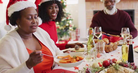 African american senior woman in santa hat serving food in plate while sitting on dining table havin