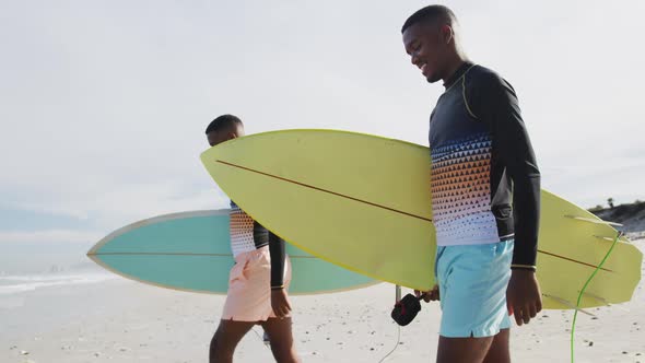 Happy african american teenage twin brothers walking on a beach carrying surfboards