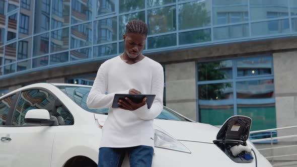 Stylish African American Man Man Stands Near an Electric Car Charging and Uses His Tablet