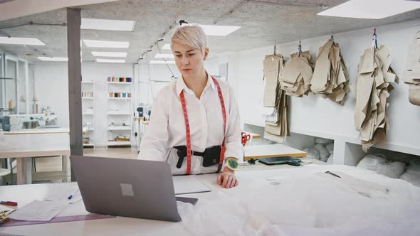 Female Tailor Working on Laptop and Writing Down Info Into Notepad Standing at Table in Sewing