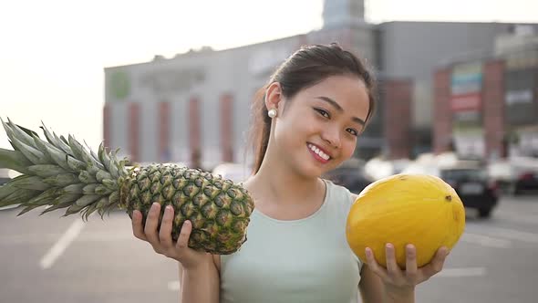 Cheerful Asian Woman Showing Melon and Peanapple in Her Hands Near Big Store