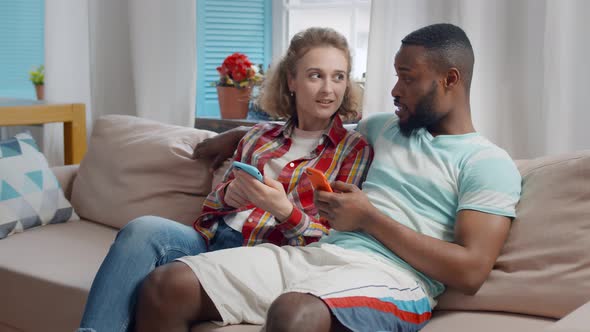 Cheerful Diverse Man an Woman in Love Using Phones Sitting on Sofa