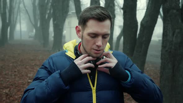 Man Athlete Puts on Wireless Headphones in a City Park, Cold Foggy Morning