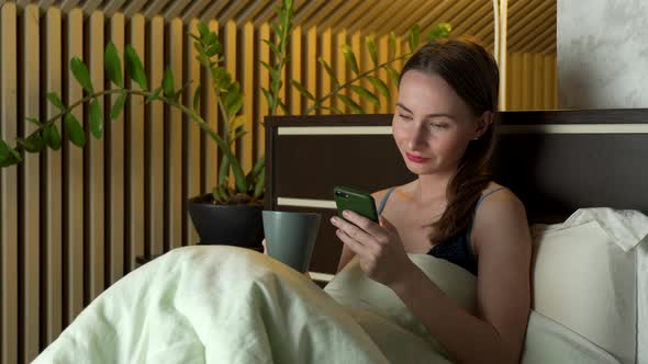 Woman Lies in Bed in the Evening and Uses a Smartphone and Drinks Tea