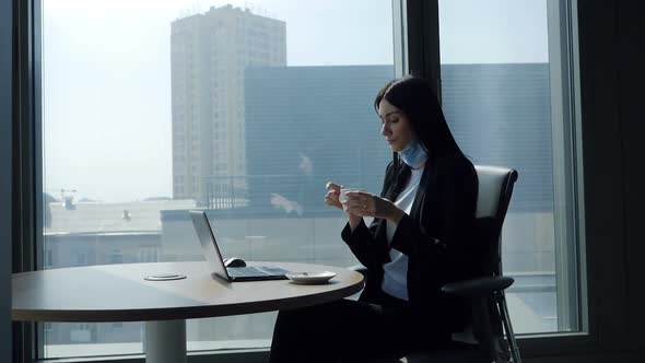 Female Worker in a Surgical Mask at the Workplace Typing on a Laptop Keyboard Working and Drinking