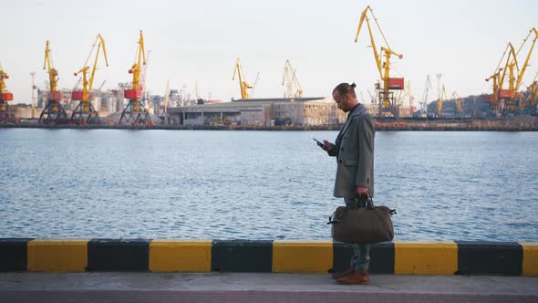 Portrait of Young Handsome Hipster Man with Bag Standing Using His Mobile Phone with Sea Port on