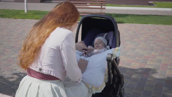 Beautiful Red-haired Woman Playing with Her Kid Lying in the Pram in the Park. The Lady Enjoying the
