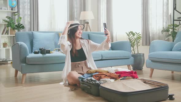 Young Woman Try Hat And Selfie While Packing Clothes In Suitcase At Home, Preparing For Vacation