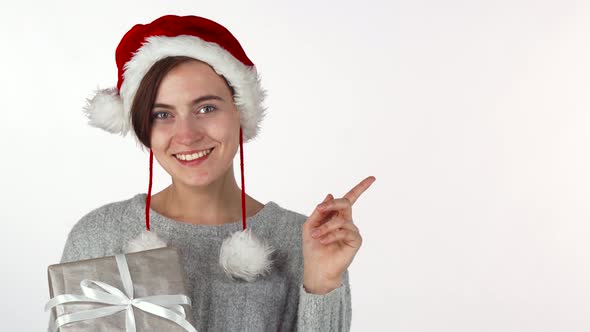 Happy Beautiful Woman in Christmas Hat Holding a Present, Pointing at Copyspace