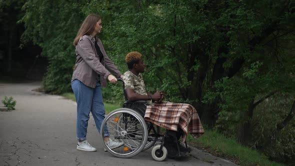 Caucasian Girlfriend Pushing Wheelchair with African American Boyfriend to Lake Pointing Talking