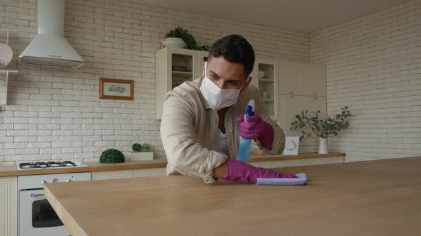A handsome man in a protective mask and gloves wipes the table with a rag and disinfectant.