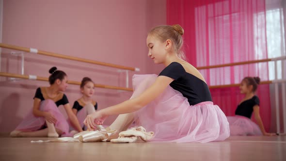 Five Ballerina Girls in Beautiful Dresses Sitting on the Floor and One of Them Changing Her Pointe