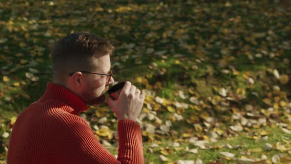 a Man in Sunglasses and an Orange Sweater Drinks Hot Tea From a Thermos While Sitting in an Autumn