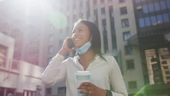 African american woman wearing face mask talking on smartphone holding coffee