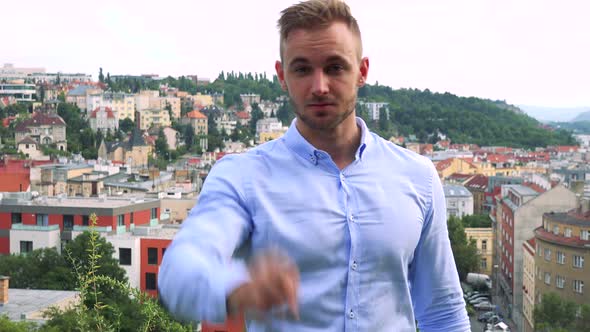 Young Handsome Business Man Points To Camera with Finger - City (Buildings) in Background