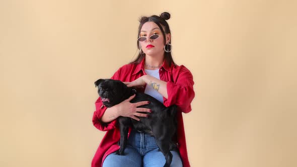 Stylish woman with tattoos strokes her black pug dog, sits with a serious face