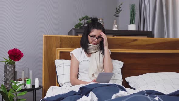 Unwell Sick Young Woman with Dark Hair which Sitting in Bed at Home and Using Tablet PC
