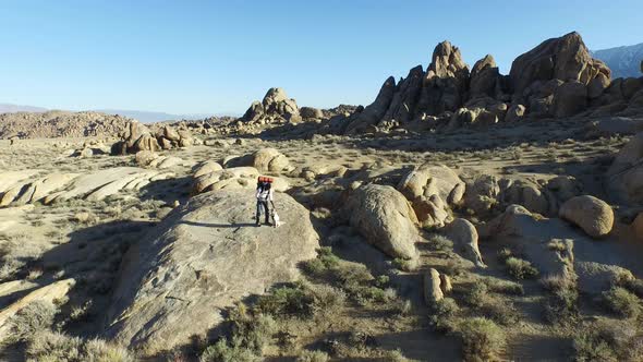 Aerial shot of a young man backpacker standing on a boulder with his dog in a desert mountain range