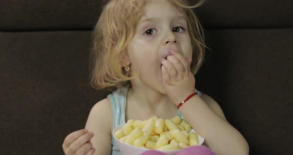 Girl Sitting on Sofa and Eating Corn Puffs. Child Smiling and Taste Puffcorns