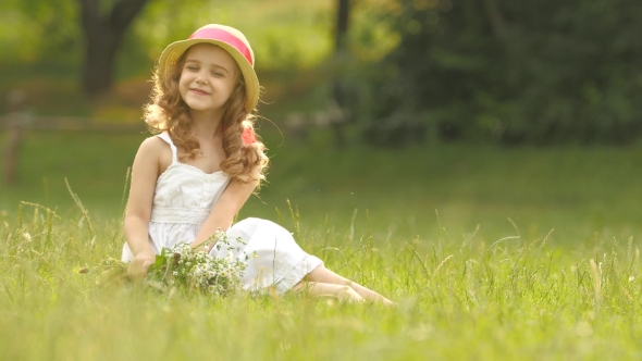 Baby Sits on a Glade in a Sarafan and Hat, Holds Wild Flowers and Smiles