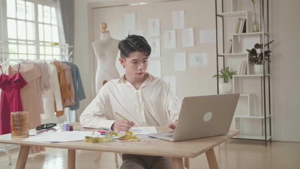 Asian Male Designer Working On Laptop And Sketching At Desk In Studio