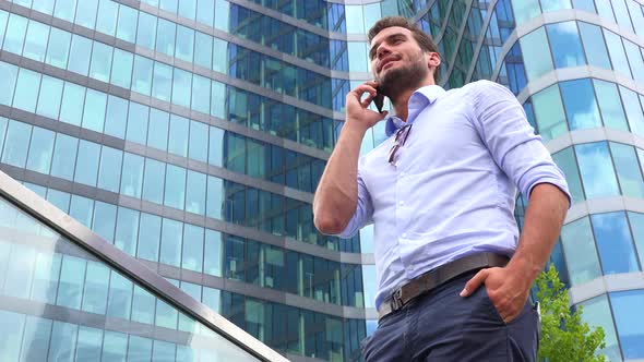 A Young Handsome Businessman Talks on a Smartphone - Closeup From Below - an Office Building