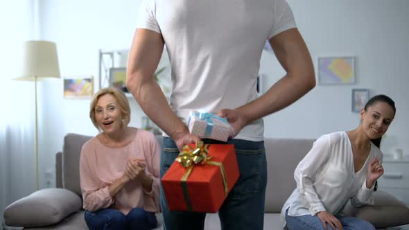 Curious Wife and Mother-In-Law Looking at Man Holding Gift Boxes Behind Back