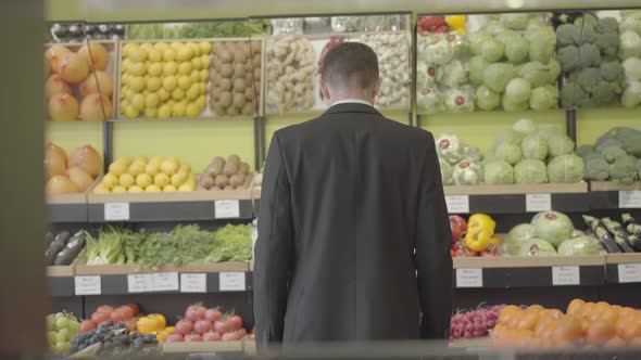 Back View of Young Caucasian Man in Suit Looking Through Shelves with Fruits and Vegetables in