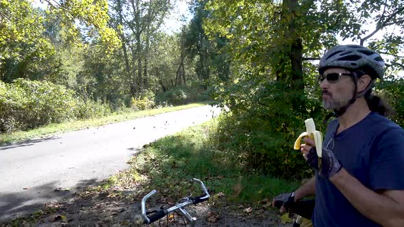 Mature man in sunglasses, helmet and gloves next to his bike, eating a banana on the side of the roa