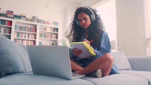 Focused Indian Woman Writes in Notebook Not Distracted on Sofa with Laptop alt