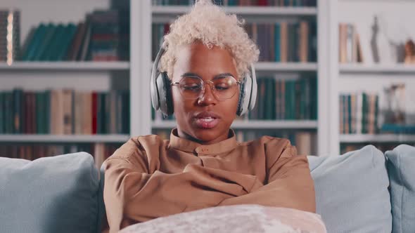 Young Tired African American Woman in Headphones Sits on Sofa in Living Room